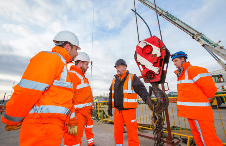 A group of construction workers stood having a conversation with a crane hook in the middle
